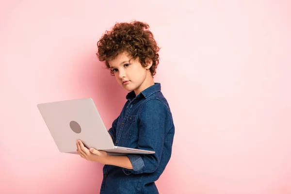 Curly boy in blue denim shirt using laptop on pink — Stock Photo