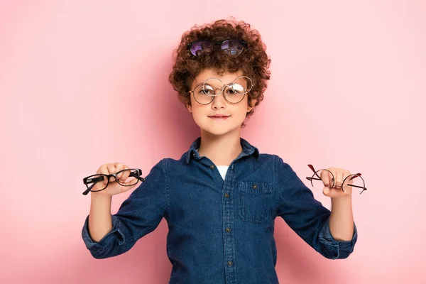 Радісний хлопчик тримає купу окулярів, дивлячись на камеру на рожевому — стокове фото