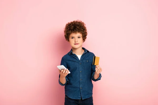 Criança encaracolado segurando smartphone e cartão de crédito enquanto compras on-line em rosa — Fotografia de Stock