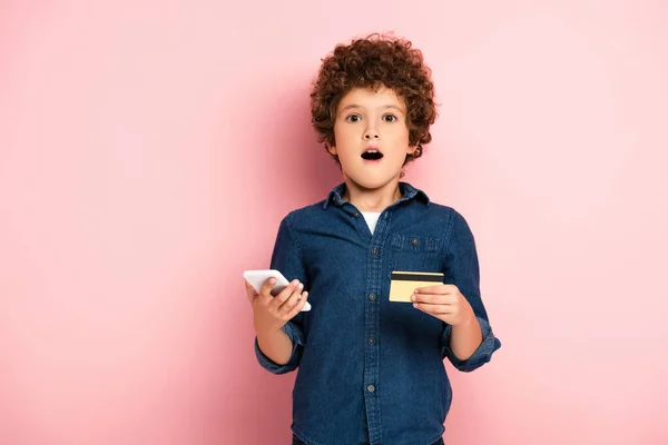 Garçon choqué tenant smartphone et carte de crédit tout en magasinant en ligne sur rose — Photo de stock