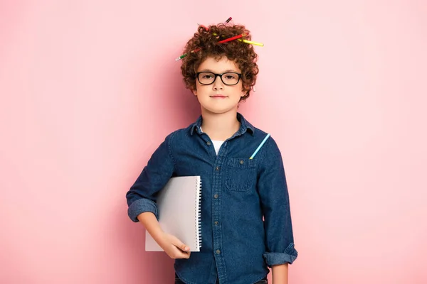 Pleased kid in glasses with color pencils in curly hair holding notepad on pink — Stock Photo