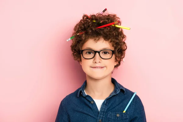 Pleased kid in glasses with color pencils in curly hair on pink — Stock Photo