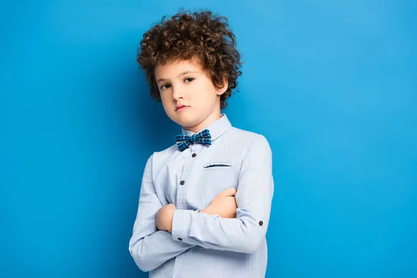 Enfant bouclé en chemise et noeud papillon debout avec les bras croisés sur bleu — Photo de stock