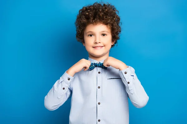 Garoto encaracolado na camisa tocando gravata arco e sorrindo no azul — Fotografia de Stock