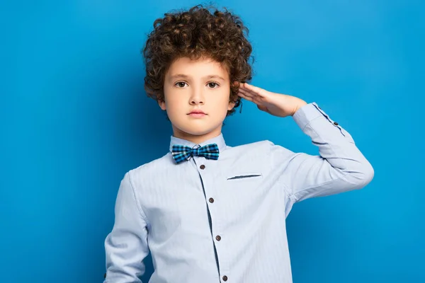 Curly kid in shirt and bow tie showing honor gesture on blue — Stock Photo
