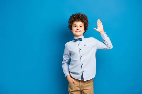 Joyful kid in shirt and bow tie standing with hand in pocket and waving hand on blue — Stock Photo