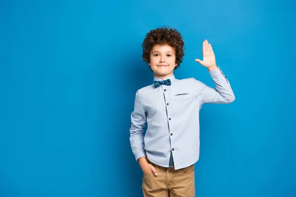 Gioioso bambino in camicia e papillon in piedi con mano in tasca e mano ondulata sul blu — Foto stock