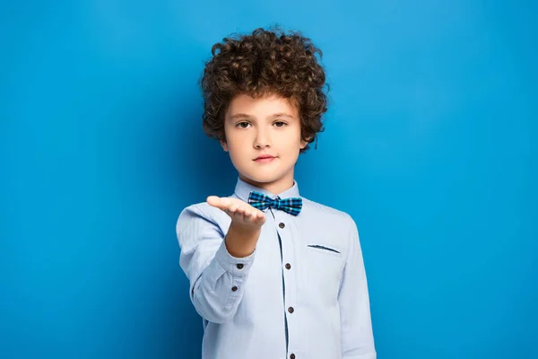 Curly kid in shirt and bow tie pointing with hand and looking at camera on blue — Stock Photo