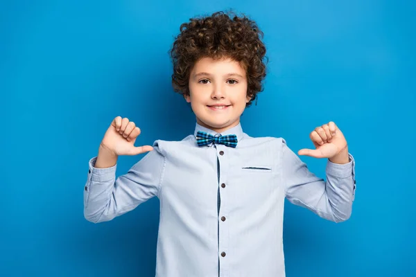 Joyful kid in shirt and bow tie pointing with fingers at himself on blue — Stock Photo