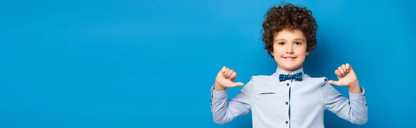 Panoramic shot of joyful kid in shirt and bow tie pointing with fingers at himself on blue — Stock Photo