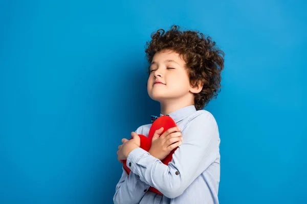 Niño alegre con los ojos cerrados sosteniendo papel rojo corazón cortado en azul - foto de stock