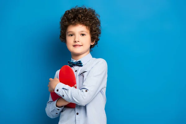 Niño alegre en camisa y pajarita sosteniendo papel rojo corazón cortado en azul - foto de stock