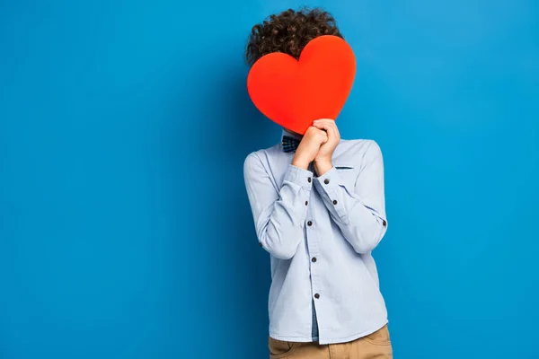 Bouclé garçon couvrant visage tout en tenant du papier rouge couper coeur sur bleu — Photo de stock