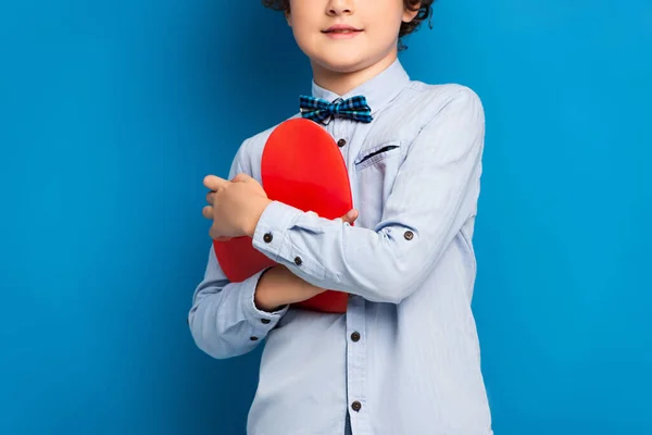 Vista ritagliata del bambino gioioso in camicia e papillon tenendo il cuore rosso tagliato carta su blu — Foto stock