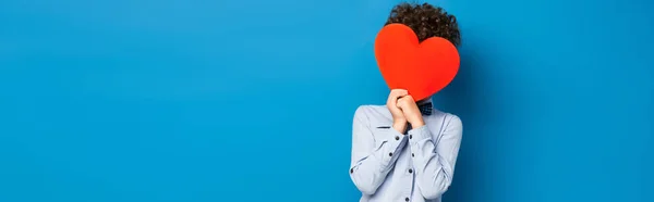 Panoramic crop of curly boy covering face while holding red paper cut heart on blue — Stock Photo
