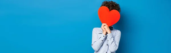 Raccolto panoramico del ragazzo riccio che copre il viso mentre tiene il cuore tagliato carta rossa su blu — Foto stock