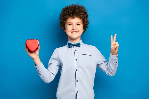 Niño complacido sosteniendo caja de forma de corazón rojo y mostrando signo de paz en azul - foto de stock