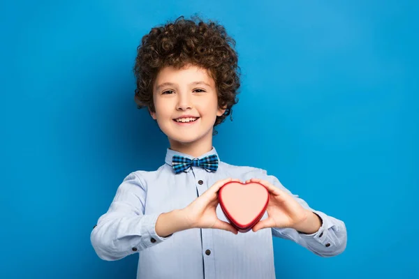 Gioioso e riccio ragazzo in possesso di scatola a forma di cuore rosso su blu — Foto stock