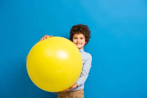 Joyful boy in shirt and bow tie holding big yellow balloon on blue — Stock Photo