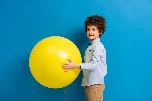 Pleased boy in shirt and bow tie holding big yellow balloon on blue — Stock Photo