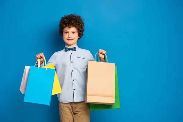 Gioioso e riccio ragazzo tenendo borse della spesa su blu — Foto stock