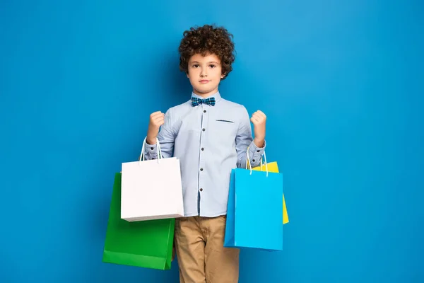 Chico rizado sosteniendo bolsas de compras y de pie en azul - foto de stock