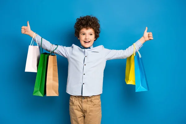 Excited and curly boy standing with outstretched hands and holding shopping bags on blue — Stock Photo