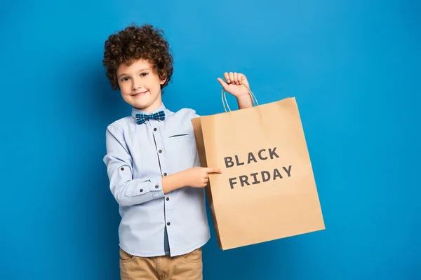 Gioioso e riccio ragazzo che punta con il dito alla shopping bag con scritta nera venerdì su blu — Foto stock