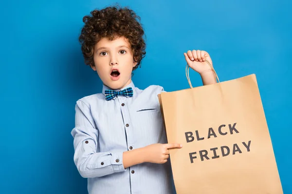 Schockierter und lockiger Junge zeigt mit dem Finger auf Einkaufstasche mit schwarzem Freitag-Schriftzug auf blau — Stockfoto