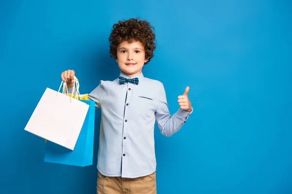 Menino alegre e encaracolado segurando sacos de compras e mostrando polegar para cima no azul — Fotografia de Stock
