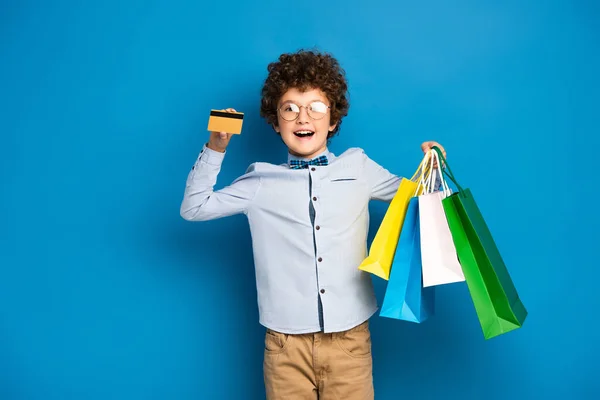 Fröhlicher und lockiger Junge in Brille mit Einkaufstaschen und Kreditkarte auf blauem Grund — Stockfoto