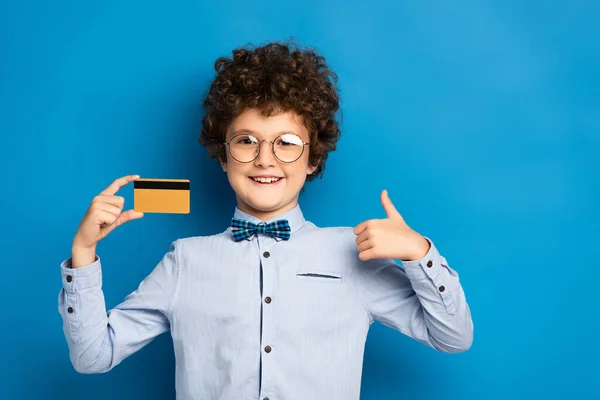 Joyful boy in glasses holding credit card and showing thumb up on blue — Stock Photo