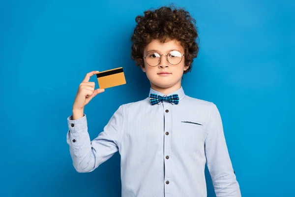 Niño rizado en gafas con tarjeta de crédito y mirando a la cámara en azul - foto de stock