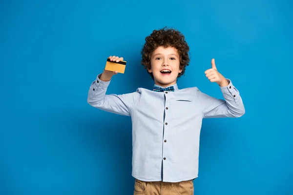 Excité garçon tenant carte de crédit et montrant pouce vers le haut sur bleu — Photo de stock