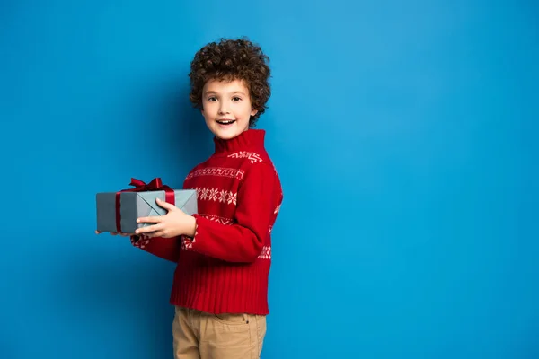 Menino animado em camisola vermelha com ornamento segurando presente no azul — Fotografia de Stock