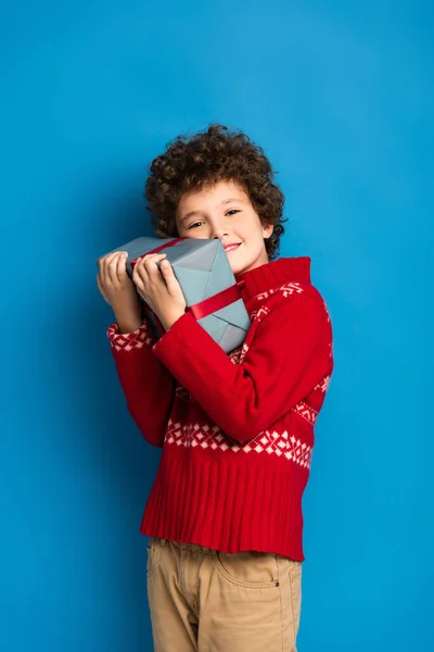 Fröhlicher Junge im roten Pullover mit Ornament umarmt Geschenk auf blau — Stockfoto