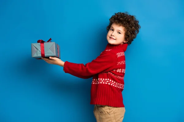 Curly boy in red sweater with ornament holding present on blue — Stock Photo