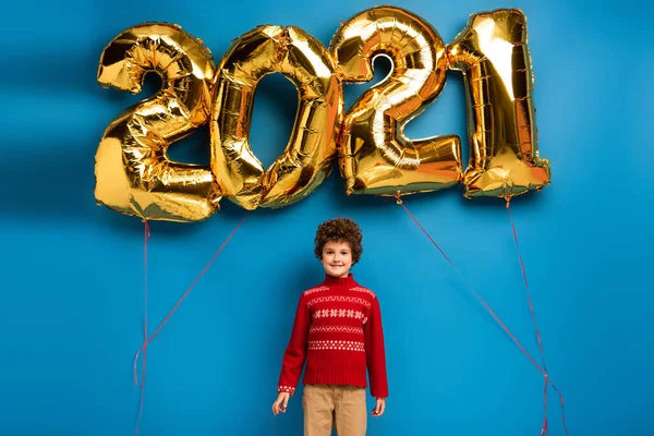 Gioioso ragazzo in maglione rosso in piedi vicino palloncini d'oro con 2021 numeri su blu — Foto stock