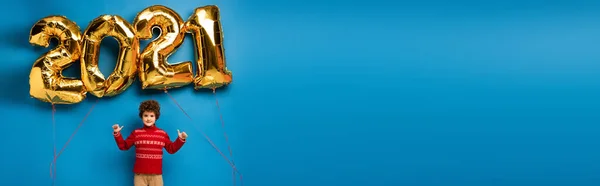 Raccolto panoramico di ragazzo riccio in maglione rosso mostrando pollici verso l'alto vicino palloncini d'oro con 2021 numeri su blu — Foto stock