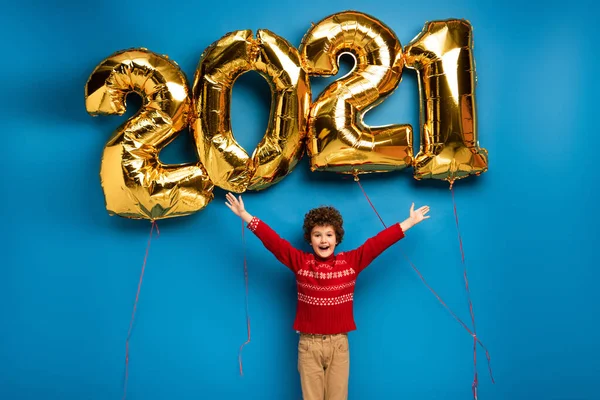 Збуджений хлопчик в червоному светрі стоїть з витягнутими руками біля золотих кульок з 2021 номерами на синьому — стокове фото