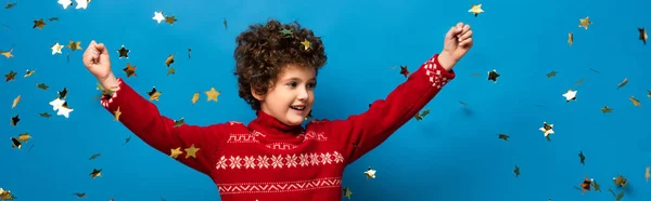 Panoramic crop of excited boy with hands above head near golden confetti on blue — Stock Photo