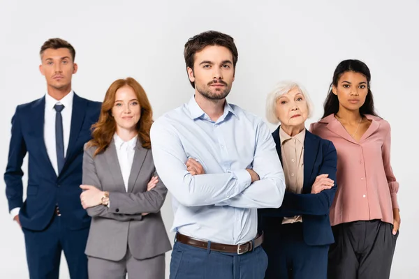 Businessman with crossed arms looking at camera near multiethnic colleagues on blurred background isolated on grey — Stock Photo