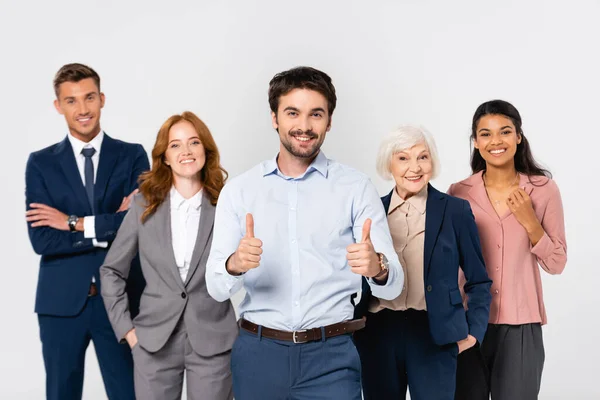 Hombre de negocios sonriente mostrando pulgares hacia arriba cerca de alegres colegas multiétnicos aislados en gris - foto de stock