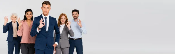 Positive multiethnic businesspeople looking at camera while showing ok gesture isolated on grey, banner - foto de stock