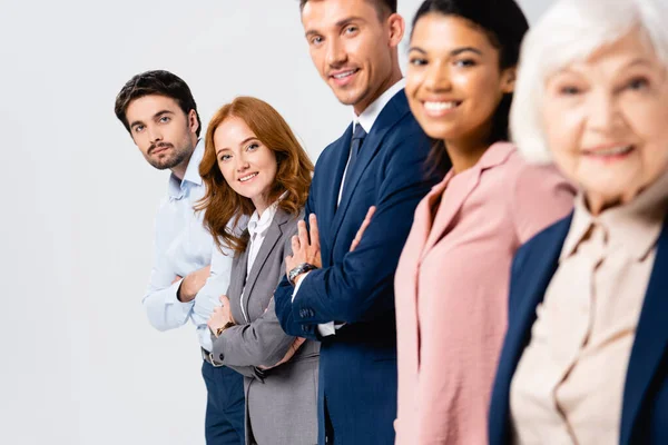 Empresarios sonrientes con los brazos cruzados mirando a la cámara cerca de colegas multiétnicos en primer plano borroso aislado en gris - foto de stock