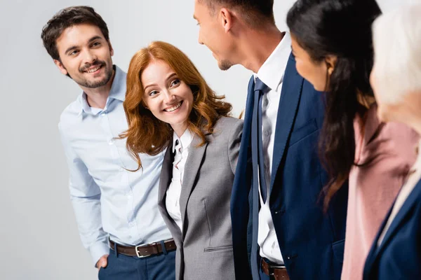 Mujer de negocios sonriente mirando a colegas multiétnicos en primer plano borroso aislado en gris - foto de stock