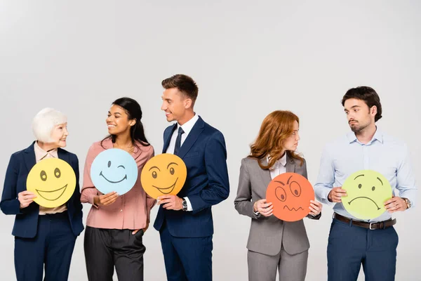 Smiling and sad multiethnic businesspeople holding cards with emotions while talking isolated on grey — Stock Photo