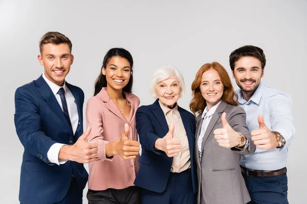 Multiethnic businesspeople showing thumbs up and smiling at camera isolated on grey — Stock Photo