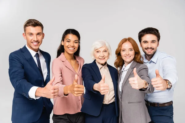 Multiethnic businesspeople showing thumbs up and smiling at camera isolated on grey — Stock Photo