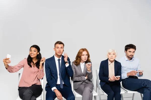 Multicultural businesspeople talking on smartphones and taking selfie on chairs isolated on grey — Stock Photo